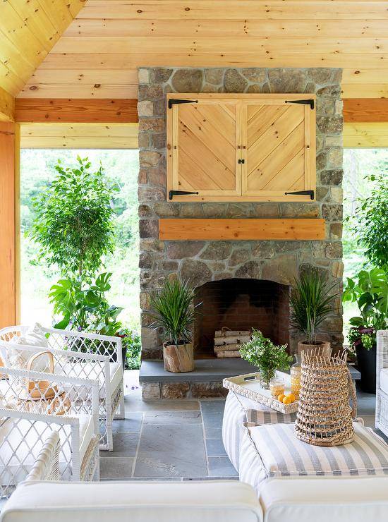 White rope chairs paired with gray stripe poufs sit on slate pavers beneath a covered patio. The patio boasts a gray stone fireplace finished with a chunky wooden mantel fixed under a TV hidden behind wood paneled doors.