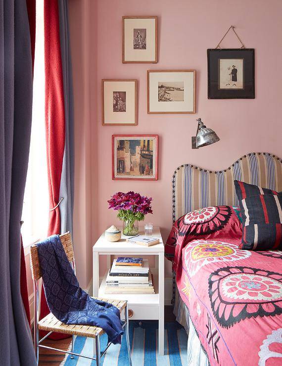 Bedroom features a beige and black striped headboard with pink bedding on pink wall, an art collage wall over a white lacquer nightstand and a blue striped rug under a woven chair with blue throw.