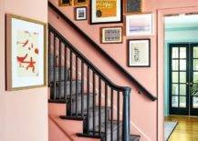An art gallery hangs from a pink wall over a wall mount black handrail complementing a black staircase covered in black runner.