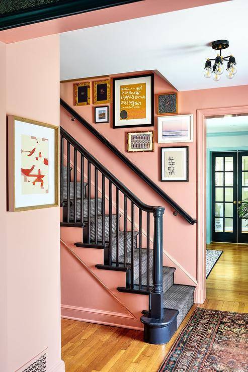 An art gallery hangs from a pink collage wall over a wall mount black handrail complementing a black staircase covered in black runner.