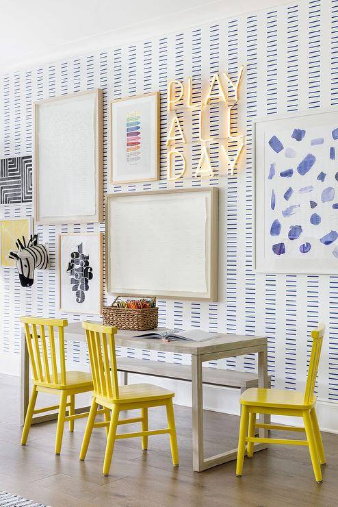 Canary yellow spindle chairs sit at a gray kid's desk under an art gallery on a blue and white wall.