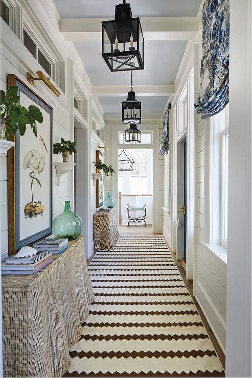 Carriage lanterns light a long cottage hallway hung from a light blue ceiling over a white and brown runner placed beneath burlap skirted console tables topped with blue glass vases. Stacked books sit on the tables beneath large flower art hung from a white tongue and groove wall beneath a brass picture light.