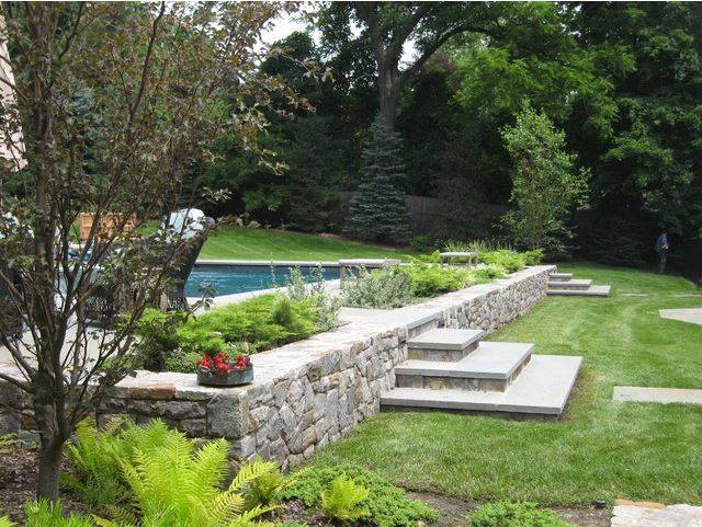 rock wall above ground pool with steps and greenery shrubs grass trees in background