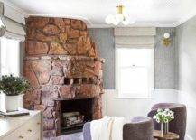 Beneath a beadboard ceiling, an eye-catching catty corner rustic stone fireplace with a matching hearth is fixed between windows dressed in gray roman shades. The windows are framed by gray fabric wallpaper lined with beadboard trim. Purple linen swivel chairs flank a gold accent table.