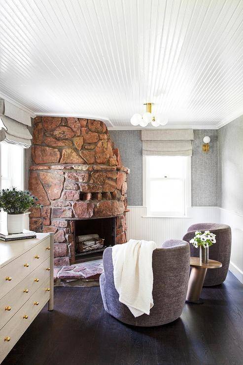 Beneath a beadboard ceiling, an eye-catching catty corner rustic stone fireplace with a matching hearth is fixed between windows dressed in gray roman shades. The windows are framed by gray fabric wallpaper lined with beadboard trim. Purple linen swivel chairs flank a gold accent table.
