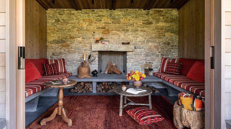 Welcoming outdoor lounge boasts a stone fireplace and a red rug framed by a u-shaped concrete bench topped with red striped cushions and red pillows.