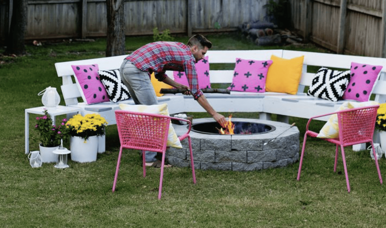 stone fire pit with benches and pink black and yellow pillows pink chairs man feeding fire with wood
