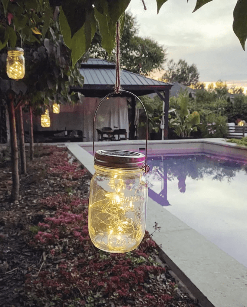 mason jar with string lights inside hanging from tree by pool