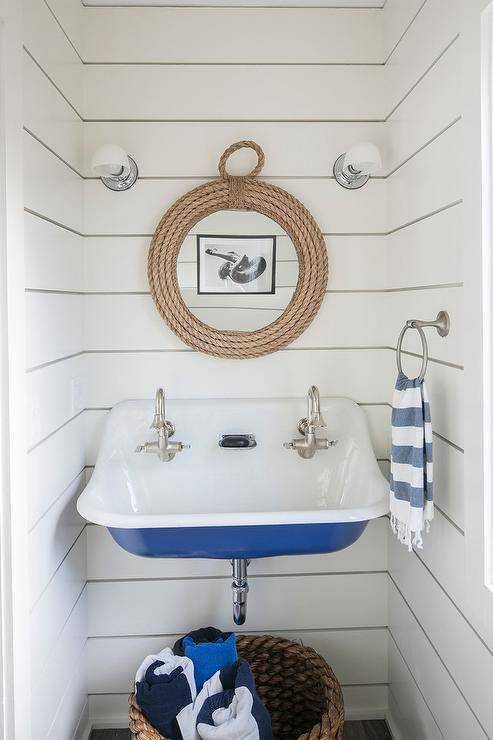 Small white and blue beach style boys' bathroom boasts a woven towel basket placed beneath a blue Kohler Brockway Sink finished with stain nickel vintage style faucets. The sink is mounted to a white tongue and groove wall under a round rope mirror hung between nickel and white glass sconces.