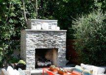 Backyard patio features a gray stone fireplace with light gray wicker outdoor sofas with a red tray outdoor tables and a blue cube stool.