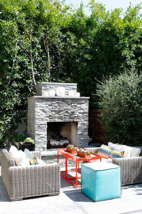 Backyard patio features a gray stone fireplace with light gray wicker outdoor sofas with a red tray outdoor tables and a blue cube stool.