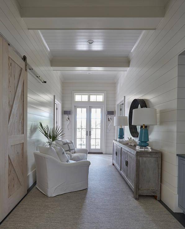 Beach style bedroom hallway is filled with a shiplap wall lined with white slipcovered chairs adorned with white and gray pillows facing a gray wash cabinet topped with blue lamps tucked under a black convex mirror.