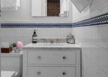 Small white bathroom features a frameless medicine cabinet mounted to a white wall finished with white grid porcelain lower wall tiles accented with blue border tiles. The medicine cabinet is located above a small white bath vanity fitted with a slatted shelf, nickel knobs, and a carrera marble countertop holding an oval sink with a polished nickel faucet. Beside the bath vanity, a toilet is positioned beneath a polished nickel towel bar.