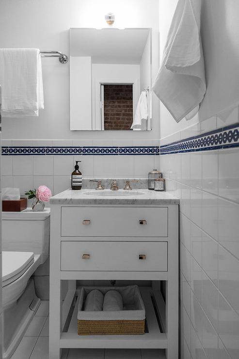 Small white bathroom features a frameless medicine cabinet mounted to a white wall finished with white grid porcelain lower wall tiles accented with blue border tiles. The medicine cabinet is located above a small white bath vanity fitted with a slatted shelf, nickel knobs, and a carrera marble countertop holding an oval sink with a polished nickel faucet. Beside the bath vanity, a toilet is positioned beneath a polished nickel towel bar.