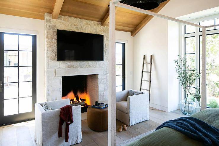 Cottage style master bedroom features a white stone fireplace fitted with a TV and positioned between a window and a glass paneled door. Natural linen club chairs sit on a wide plank wood floor and face a round brown leather pouf.