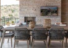 Covered patio features dark gray rope outdoor chairs with a brown teak outdoor table and a tv mounted on an outdoor stone fireplace.