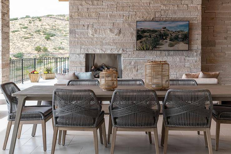 Covered patio features dark gray rope outdoor chairs with a brown teak outdoor table and a tv mounted on an outdoor stone fireplace.
