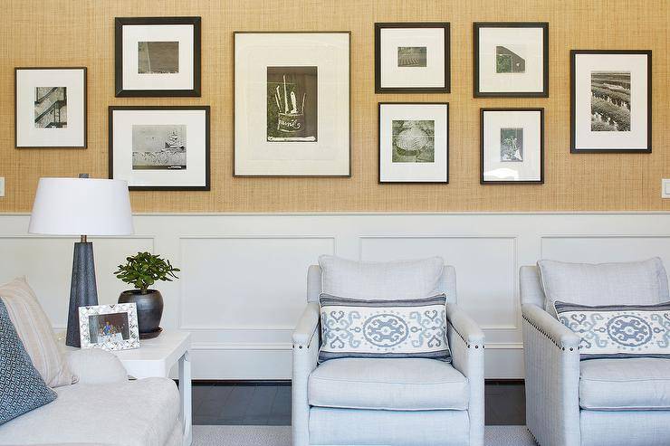 Gallery wall displayed on a gold Raffia wallpaper wall above wainscoting trim in a living room furnished with light blue accent chairs and blue Ikat lumbar pillows