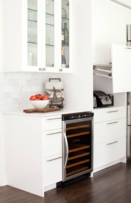 Stunning white kitchen with glass front upper cabinets lined with glass shelves over a marble subway tiled backsplash alongside a white counter above white flat front base cabinets accented with brushed nickel pulls. The drawers frame a glass front wine fridge which stands to the left of a small appliances cabinet with upward opening cabinet door.