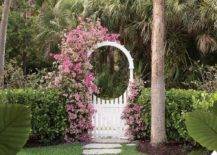Climbing pink flowers accent a white trellis fence gate opening to concrete step pavers surrounded by grass.
