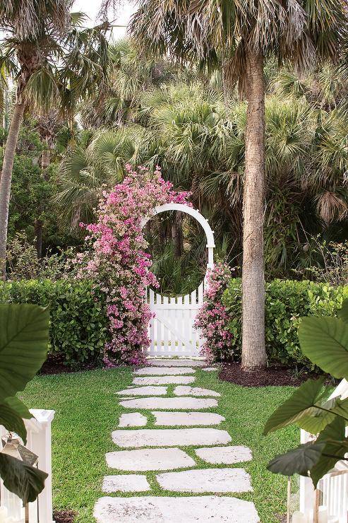 Climbing pink flowers accent a white trellis fence gate opening to concrete step pavers surrounded by grass.
