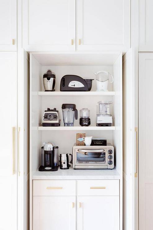 Folding doors conceal a cabinet holding small kitchen appliances on white shelves over white cabinets adorned with antique brass hardware.