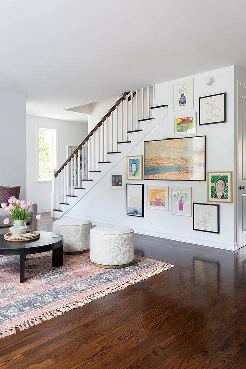 Living room features an eclectic art gallery on a staircase wall and a round black coffee table with ivory boucle stools on a pink and black rug.