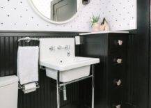 White and black contemporary bathroom is clad in white and black penny floor tiles positioned beneath a washstand boasting chrome legs and a white porcelain sink paired with a chrome backsplash mounted faucet kit. The sink vanity is located in front black beadboard trim, beside a chrome towel ring and beneath a round white mirror hung from an upper wall covered in eye pattern wallpaper. A small black cabinet sits beside the sink vanity.