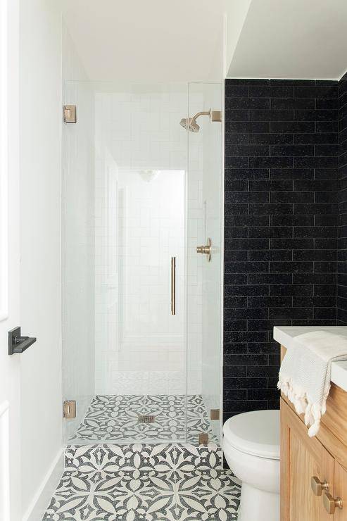 White and black mosaic floor tiles lead past a wall clad in black stacked tiles to a small walk-in shower boasting a frameless glass door and a brushed gold shower kit fixed to white subway staircase pattern surround tiles.