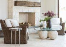 Brown wicker chairs accented with white cushions sit on ivory travertine tiles on either side of a glass top coffee table placed in front of an ivory stone fireplace finished with an ivory stone herringbone fire box and a rustic wood mantel.