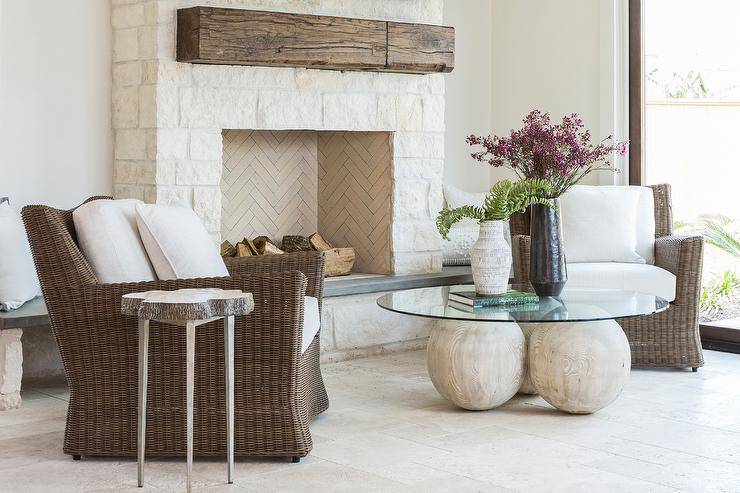 Brown wicker chairs accented with white cushions sit on ivory travertine tiles on either side of a glass top coffee table placed in front of an ivory stone fireplace finished with an ivory stone herringbone fire box and a rustic wood mantel.