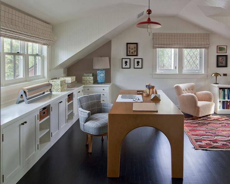 Beautifully designed home office boasts a red lantern hung over a brown wooden desk matched with a gray chair placed on an ebony stained wood floor. Behind the desk, white built-in cabinets and shelves are fixed under a window.