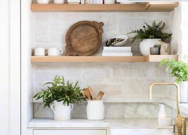 Kitchen features blond floating shelves on a rustic stone backsplash and white cabinetry accented with bronze pulls.