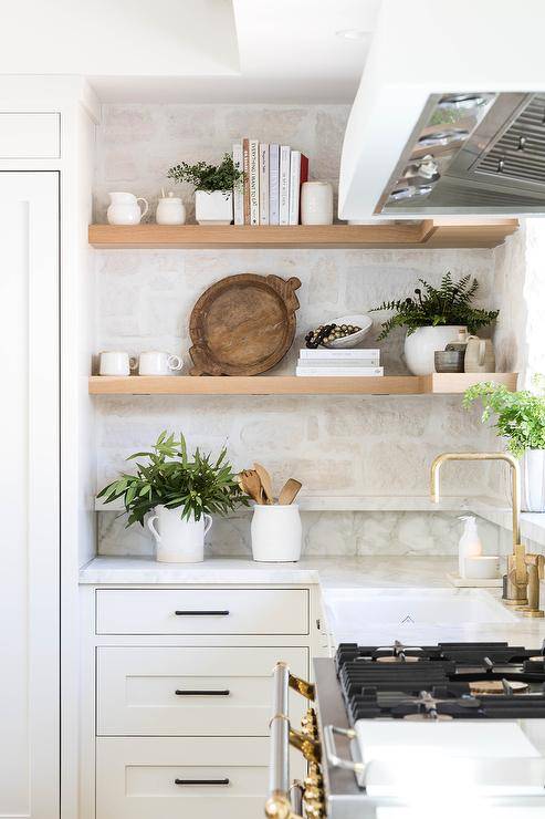 Kitchen features blond floating shelves on a rustic stone backsplash and white cabinetry accented with bronze pulls.