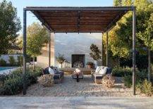 Wood and iron pergola with a fireplace in a backyard furnished with two brown teak outdoor sofas on a gravel surface.