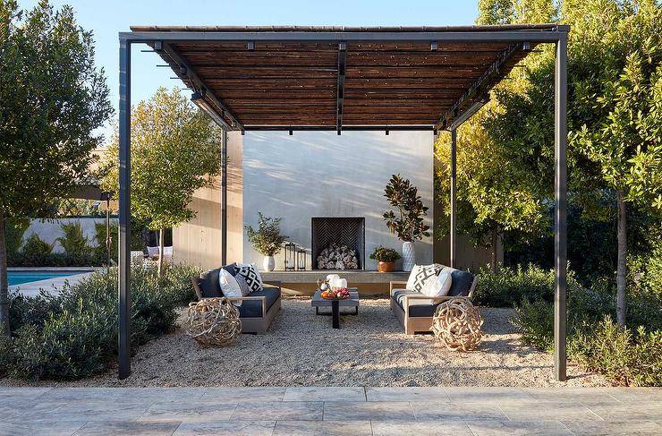 Wood and iron pergola with a fireplace in a backyard furnished with two brown teak outdoor sofas on a gravel surface.
