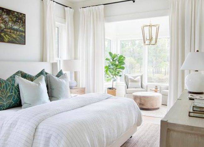 Welcoming master bedroom sitting area features a white curtain hung from an oil rubbed bronze rod in front of a nook boasting a round ottoman placed in front of dove gray wingback chairs lit by a brass lantern hung in front of windows.