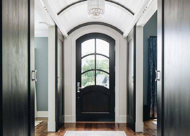 Long hallway with a plank barrel ceiling elegantly displaying a cascading chandelier over a light gray vintage rug layered on oak floors. A black arch front door brings a unique look contrary to a dull standard door.