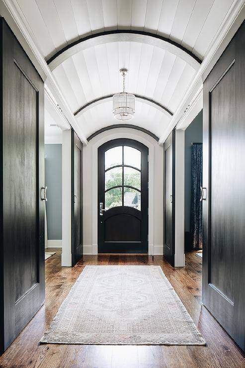 Long hallway with a plank barrel ceiling elegantly displaying a cascading chandelier over a light gray vintage rug layered on oak floors. A black arch front door brings a unique look contrary to a dull standard door.