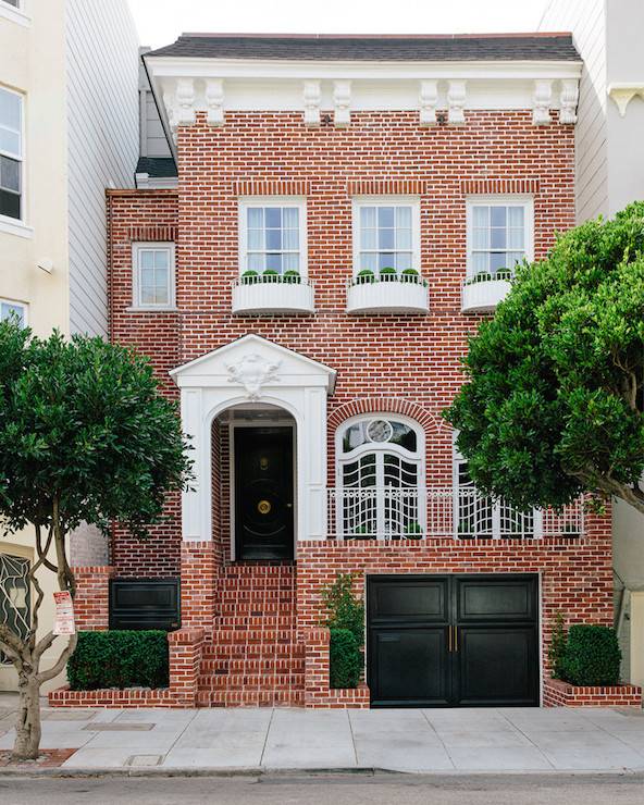 Red brick home is accented with glossy black door situated next to garage located below first level of home.