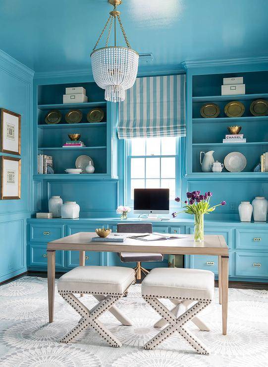 Turquoise blue home office features white x-stools placed on a gray rug at a gray French desk matched with a dark gray velvet task chair lit by a white beaded chandelier hung from a turquoise blue painted ceiling. Behind the desk, turquoise blue built-in cabinets are fixed against a turquoise blue wall and under turquoise blue cabinets flanking a window covered in a turquoise blue striped roman shade.