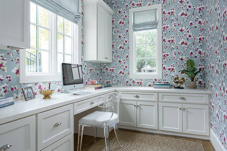 Blue and pink wallpaper accents a home office boasting an acrylic chair placed on a diamond pattern jute rug at a white l-shaped built-in desk fitted with polished nickel cup pulls and a white quartz countertop. Above the desk, a window dressed in a blue roman shade is flanked by white shaker cabinets.