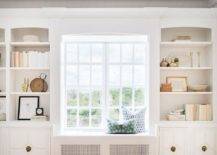 White styled shelves fixed over white cabinets with brass hardware flank a built-in window seat.