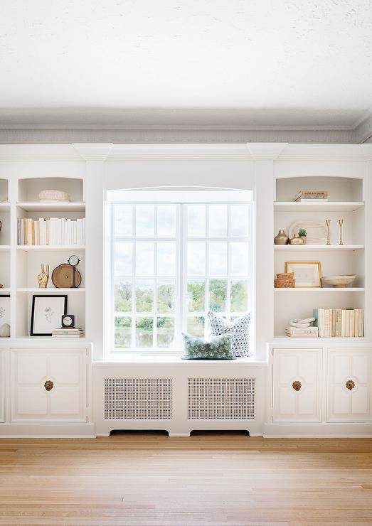 White styled shelves fixed over white cabinets with brass hardware flank a built-in window seat.