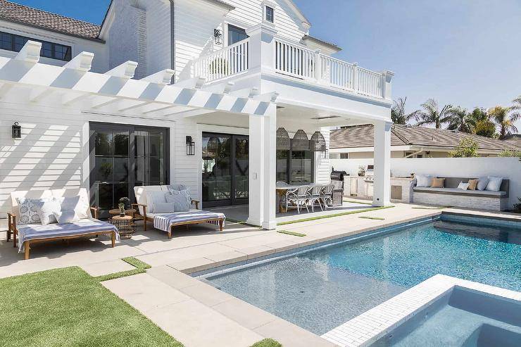 Teak double pool loungers with blue stripe throw blankets sit under a white pergola in front of an inground pool.