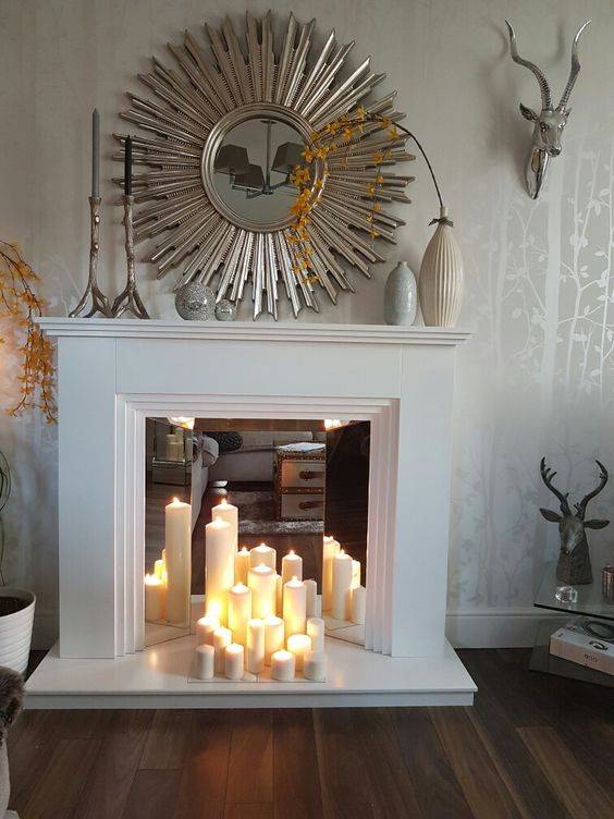 a faux white fireplace with a mirror screen and pillar candles, a sunburst mirror, vases on the mantel for decor