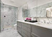 White and gray bathroom boasts marble everywhere you look! A gray and white marble walk in shower is enclosed by tall glass doors with stainless steel handles continuing on the the floors and backsplash walls. A dual gray washstand topped with white marble counters displays a black Chanel tray under a set of frameless medicine cabinets mounted side by side.