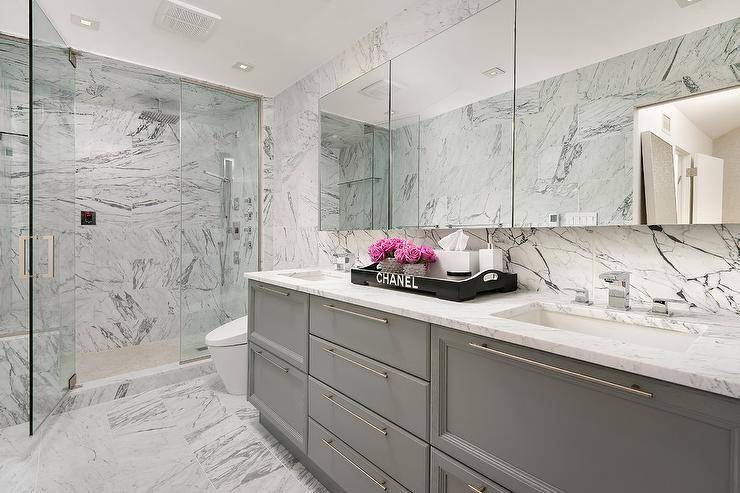 White and gray bathroom boasts marble everywhere you look! A gray and white marble walk in shower is enclosed by tall glass doors with stainless steel handles continuing on the the floors and backsplash walls. A dual gray washstand topped with white marble counters displays a black Chanel tray under a set of frameless medicine cabinets mounted side by side.