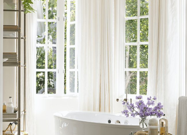 Bathroom features a bay window with oval bathtub, a round nickel and glass accent table and cream curtains.