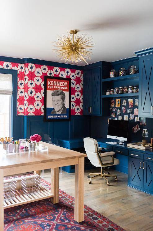 Blue home office features blue board and batten walls accented with red and blue wallpaper lined with blue crown moldings. The room is lit by a brass sputnik chandelier hung behind a blue built-in desk seating a white and gold task chair, while blue floating shelves are stacked between blue x-front cabinets.